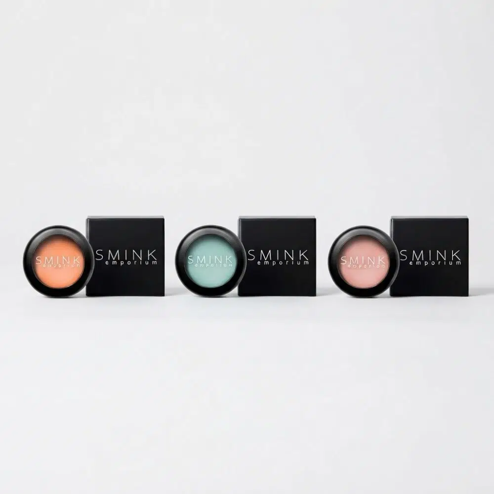 Row of four circular makeup pots with pastel shades (peach, mint, teal, pink) beside black SMINK Emporium boxes, arranged in a line on a white background.