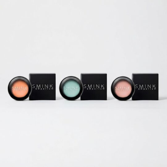 Row of four circular makeup pots with pastel shades (peach, mint, teal, pink) beside black SMINK Emporium boxes, arranged in a line on a white background.