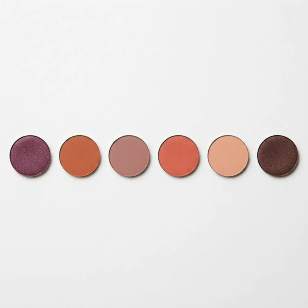 Row of six round makeup color swatches: purple, orange, taupe, coral, peach, and dark brown/purple tones on a white background.