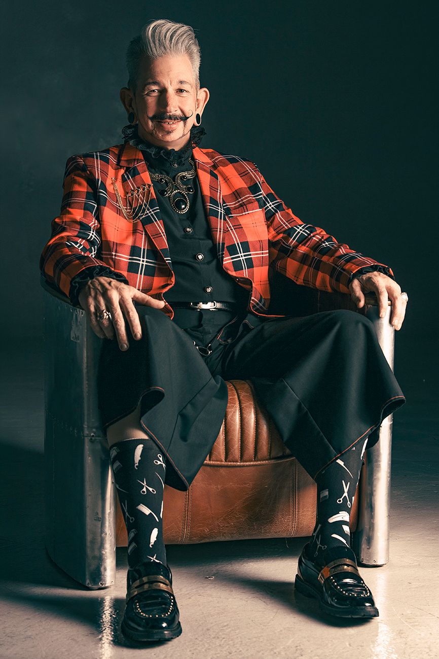 Portrait of a stylish person with a gray pompadour and mustache, wearing a red plaid blazer, seated in a vintage metal chair.