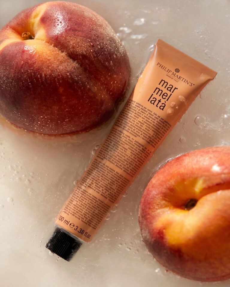 Two peaches surround a peach marmellata tube, resting in a wet sink or shallow water surface, suggesting peach jam.